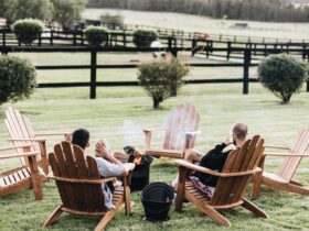 two men sitting on wood chairs on a farm by an outdoor fire pit with drinks in hand