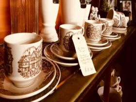 Row of 5 cups and saucers engraved with an old farm house, sitting on a wooden bench.