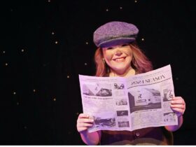 A young person in a cap, smiling and holding a newspaper.