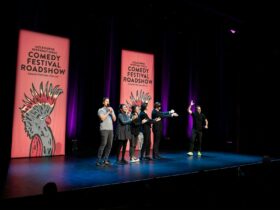 Image of 6 comedians standing on a stage in front of large banners