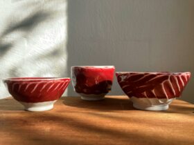Three small red bowls on a wooden shelf in front of a white wall. The light is shining from the side