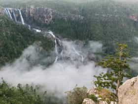 Water falls over rocky gorge, covered in cloud and mist surrounded by forest.