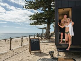 A swimwear clad female and male stand at the exit of the mobile sauna looking out to the ocean.