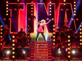 Actress dressed as Tina Turner standing in power pose singing alongside band with lights behind her