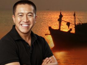 A man in black shirt is smiling directly at the camera, behind him is the sea and a fishing boat