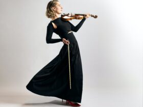Photo of a violinist in a black dress, slightly leaning backwards, against a solid grey backdrop.