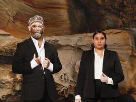 Two Aboriginal people in suits with ochre on their face stare directly to camera.