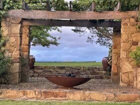A stone and wooden arbour frames a green paddock. A fire pit sits at the front.