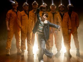 Billy Elliot dances in front of miners in shadow with headlamps on.