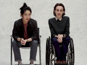 Two individuals sit next to each other against a grey background, one in a wheelchair
