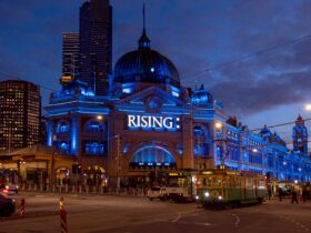 A tram passing in front of Flinders Street Station at night, with blue lights and a neon RISING sign