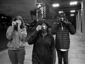 Black and white photo. Three youth photographed with digital cameras held up to their face.