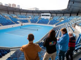 A group of four people are looking at a blue tennis court from the stands. One is a tour guide.