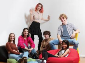 A group of six smiling young people aged 12–18 sit on bean-bags and chairs, facing the viewer.