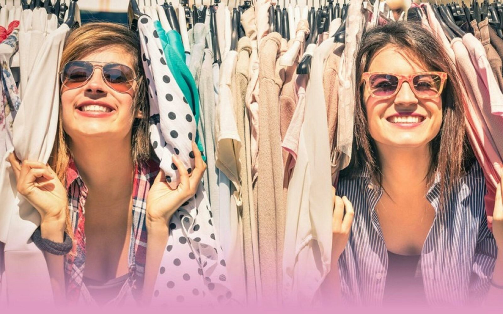 Two women wearing sunglasses and smiling while poking their heads through a rack of clothing