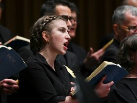 A young woman holds a printed musical score and sings passionately surrounded by a choir