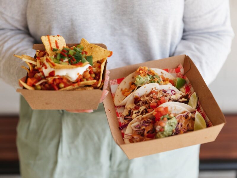 person holding two takeaway containers of food, one with tacos one with nachos