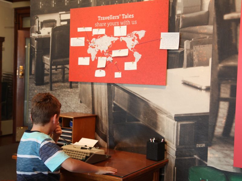 Boy thinking at typewriter in front of red and white world map showing visitor’s travel stories