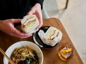 Hands holding a fluffy katsu sandwich with a bowl or ramen and a drink nearby.