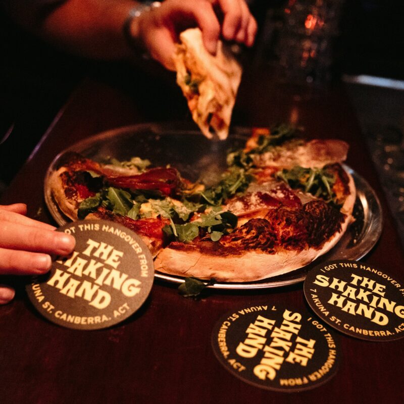 People grabbing at a pizza. The Shaking Hand branded coasters surrounding pizza