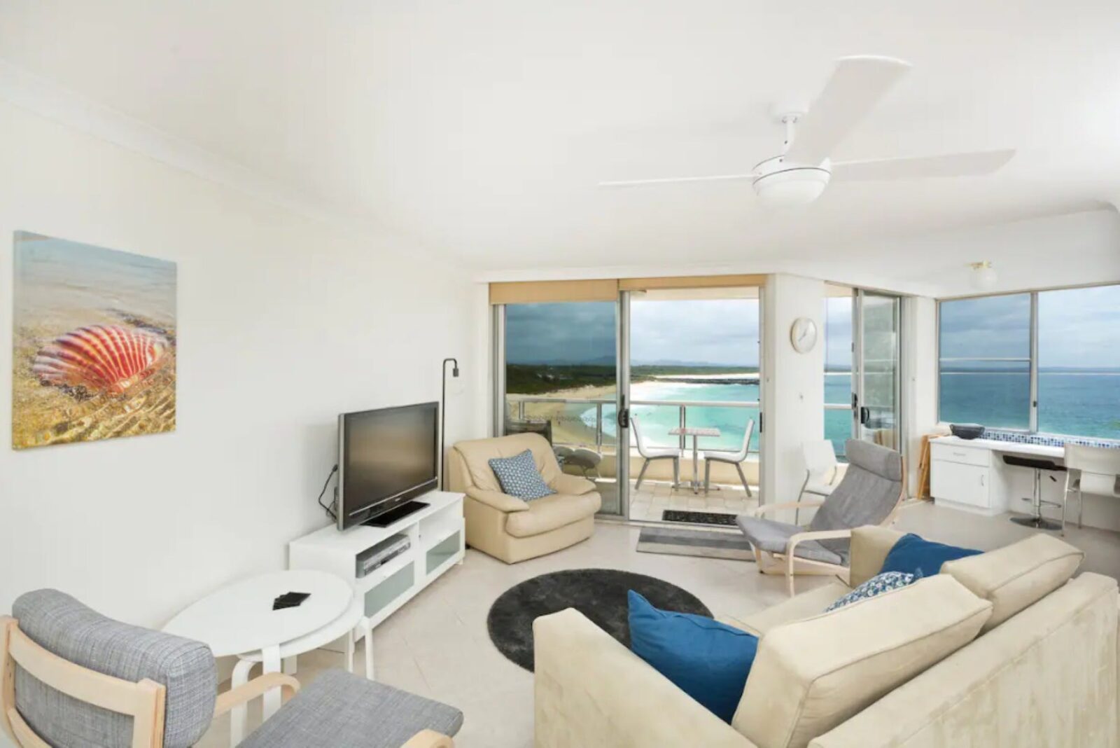Living room with lounges, ceiling fan, TV, ocean views, leading out to balcony with 2 seater setting