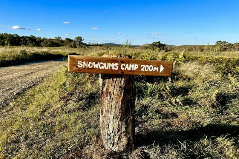Rustic timber signpost in grassy field next to dirt road, reads 'Snowgums Camp 200m'. Blue sky.