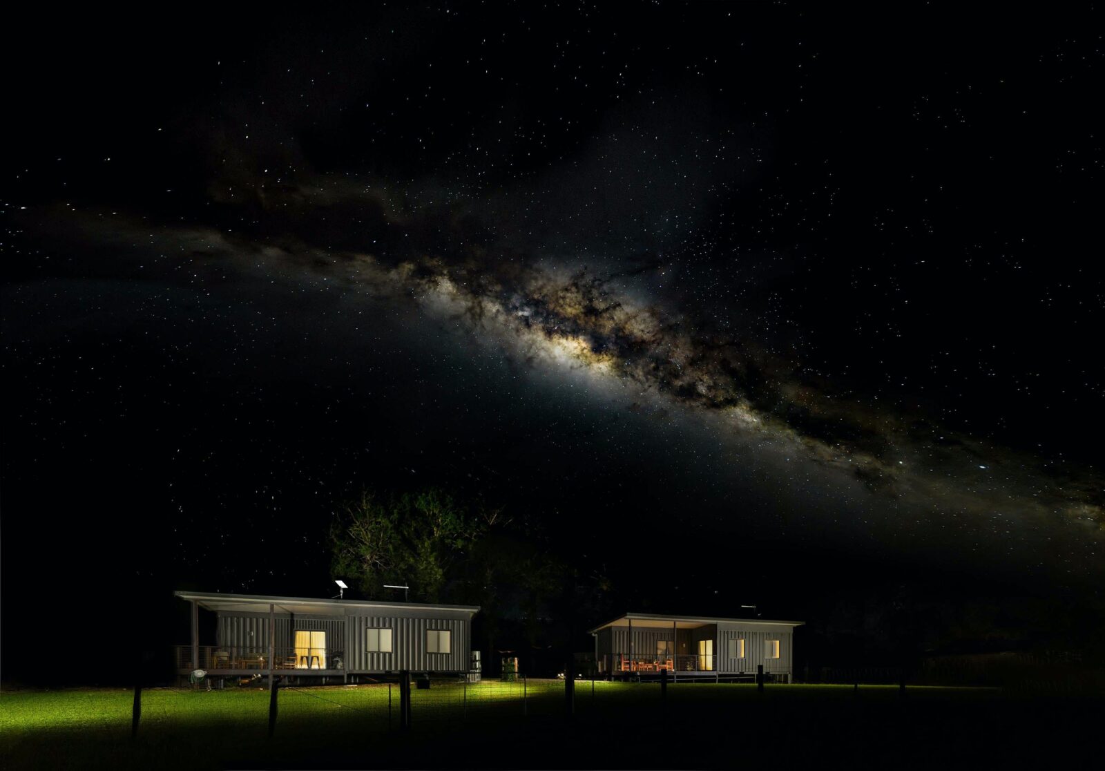 An image of both cottages sitting in a paddock at night with the Milky Way arching over the top.
