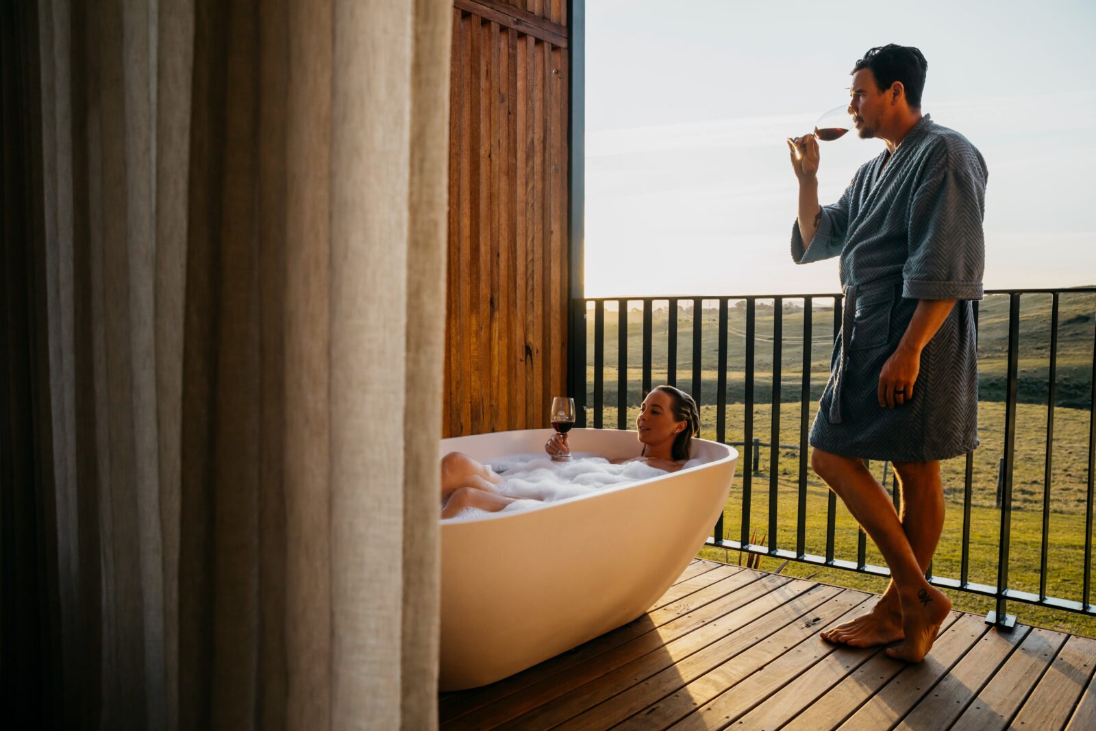 Woman enjoys outdoor bathtub drinking wine, man looking at the view drinking red wine.