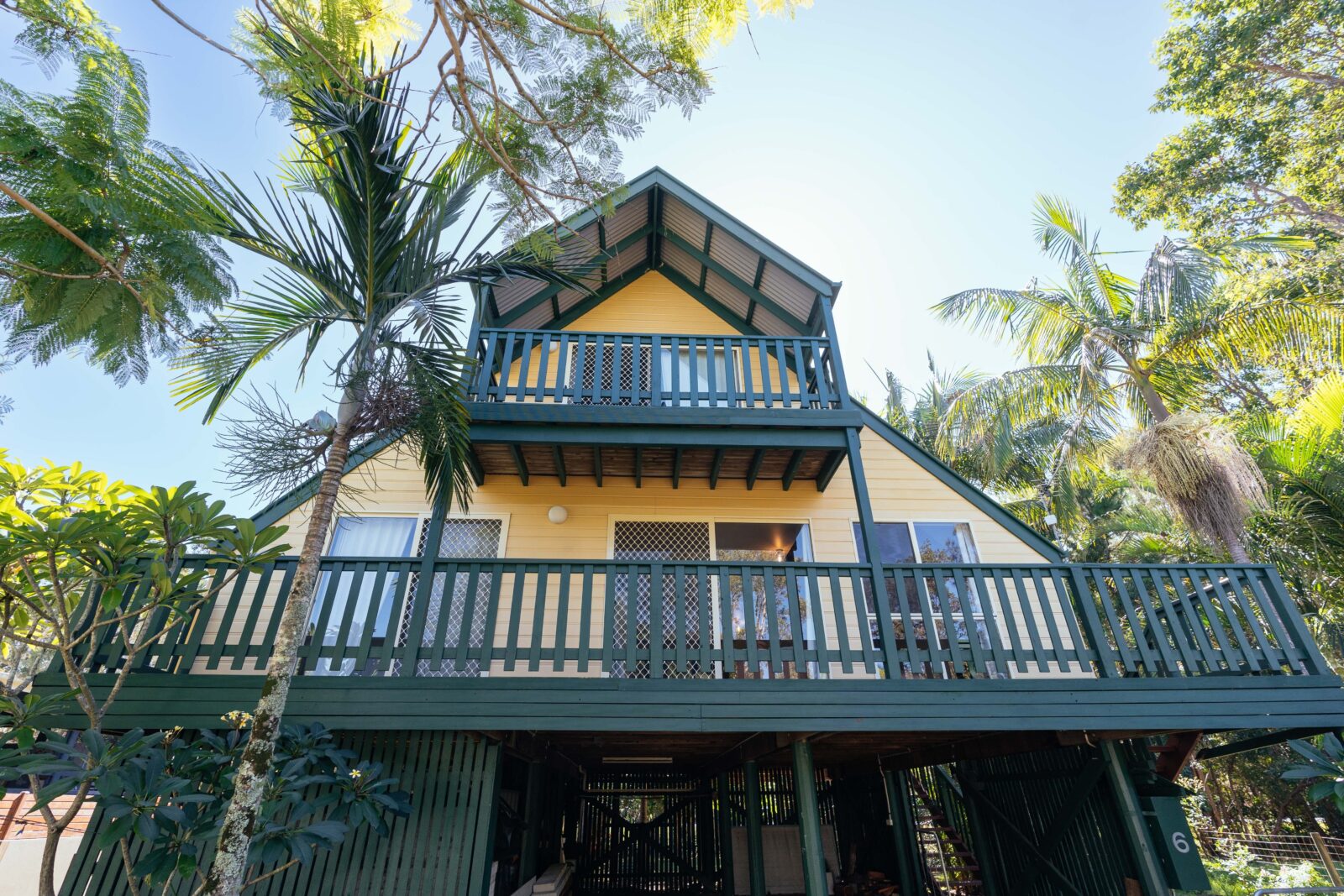 A yellow A-Frame house with green trim sits amongst tall palm trees with a blue sky