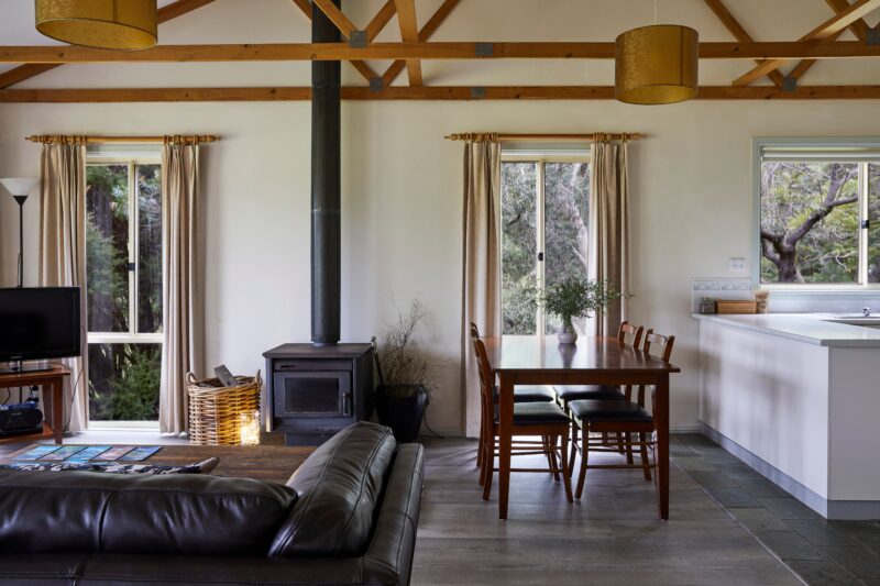 Cottage interior shows wood burner, sofa, dining table and edge of kitchen surface. Natural light