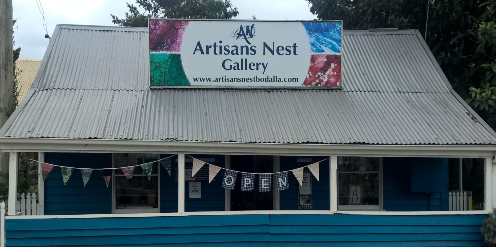 Artisans Nest Gallery exterior from highway, marine blue fence,showing roof sign and "open" Bunting