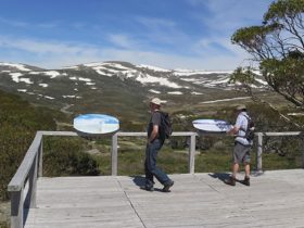 2 bushwalkers read park information at Charlotte Pass lookout, with mountains in the background.