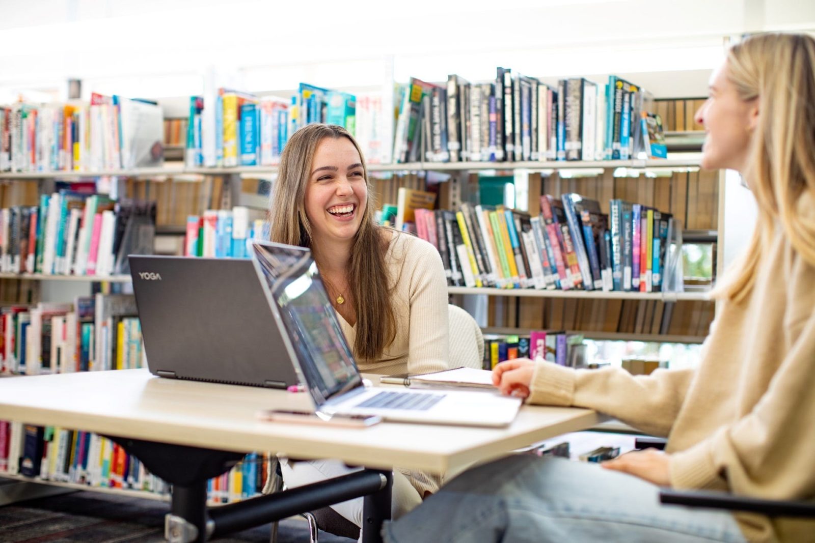 Two young women laugh while working on laptops in the library