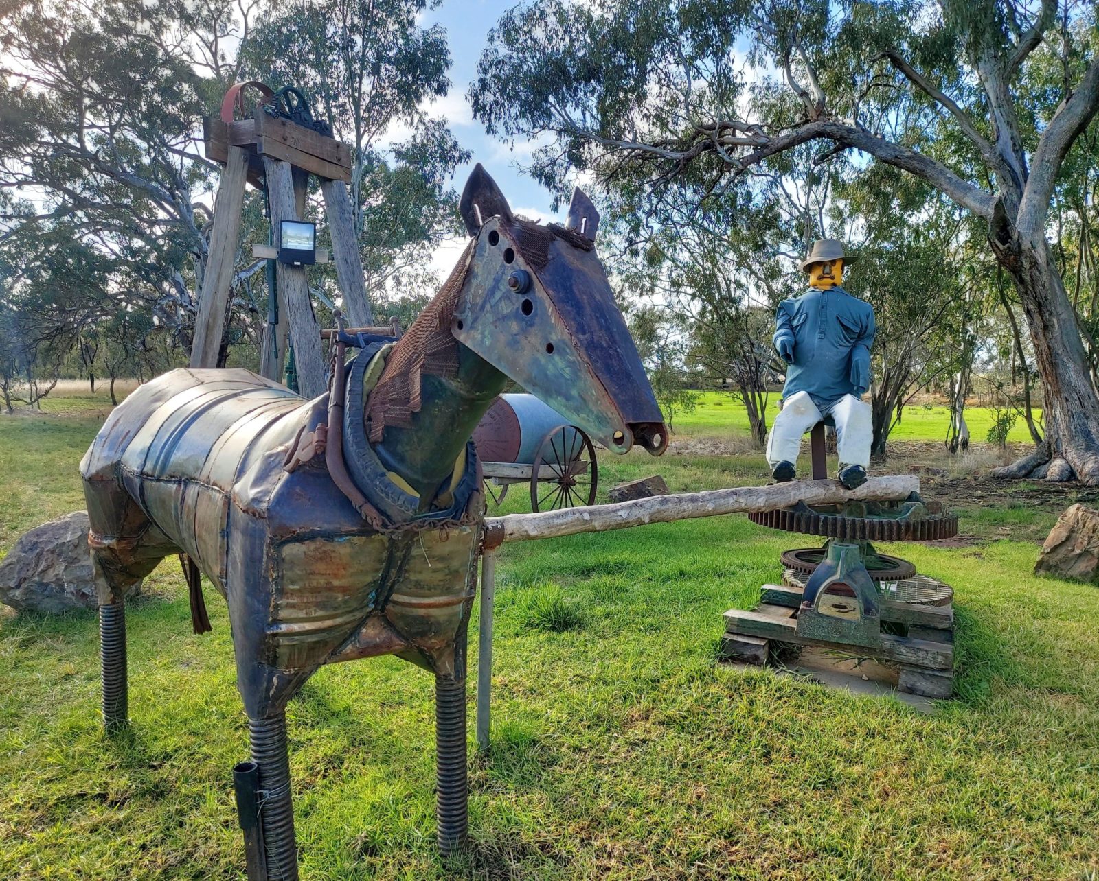 A metal horse sculpture and a man sitting at a water pump in a grassy park.