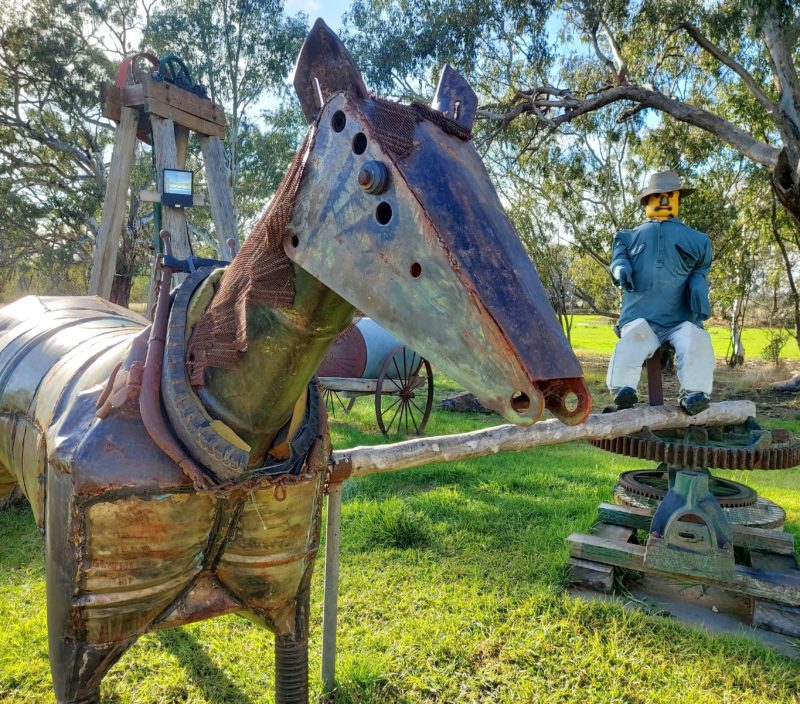 A closeup of a metal horse sculpture with a sculpture of a man at a pump in the background.
