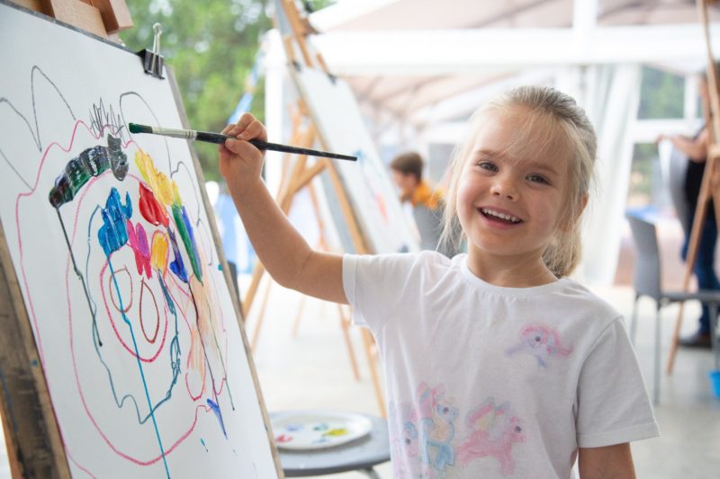 A young blond girl smiling while painting on paper on an easel.