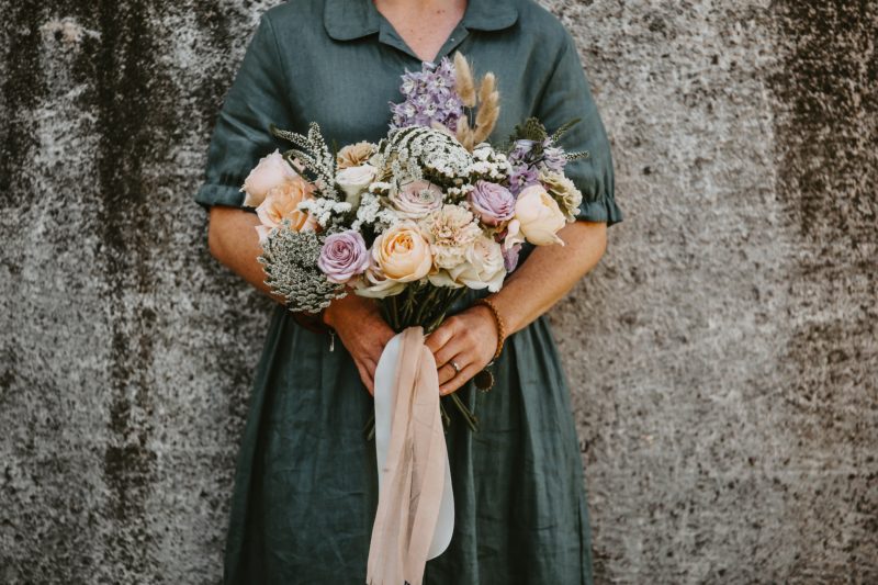 Neck down photo of a lady in a green dress holding a bouquet of pink and purple flowers with ribbons