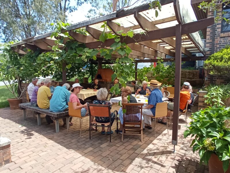 20 members sit under the shade of the awning in the Members' Courtyard for a summer lunch