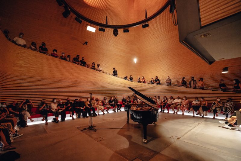 A grand piano sits in a large performance space as people gather