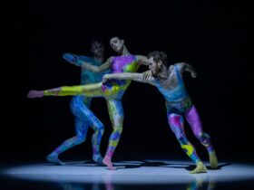 Three Sydney Dance Company dancers wearing brightly coloured body suits as they dance on stage