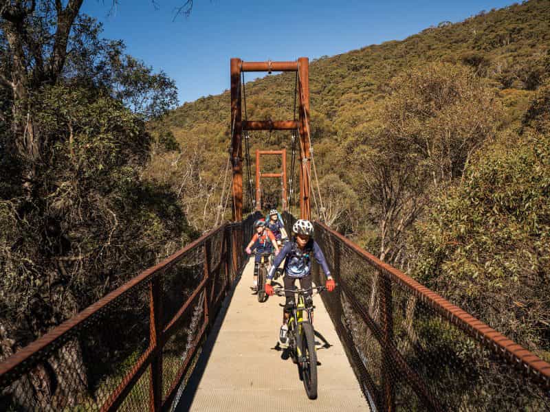 A family bike rides across a suspension bridge along Thredbo Valley track, Kosciuszko National Park