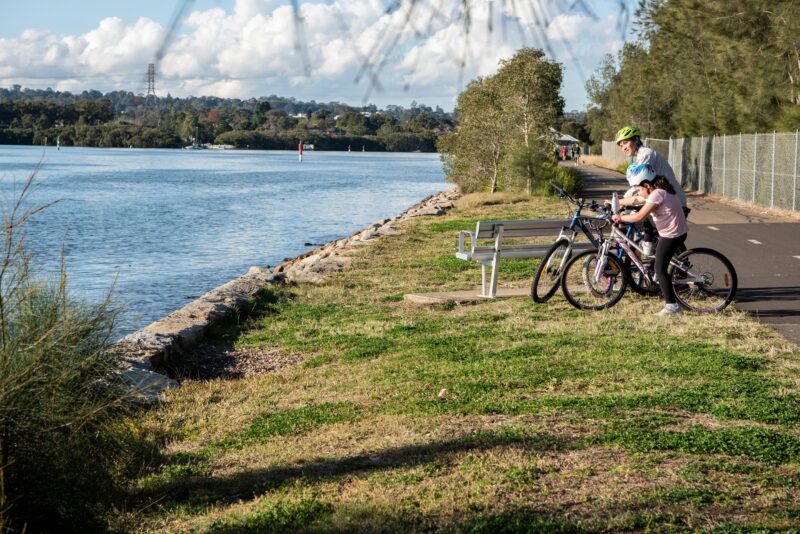 3 bike riders stopped by the river to look at the view, along a grass edge and pathway.