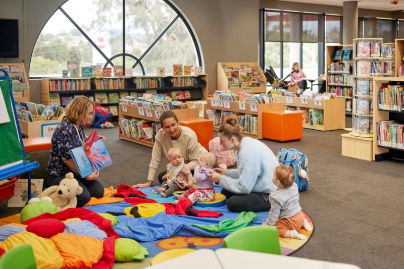 A librarian smiles and reads a book to a small group of babies and mums.