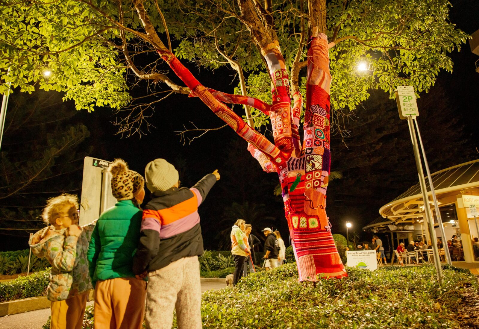 Children pointing a tree that has been yarn bombed