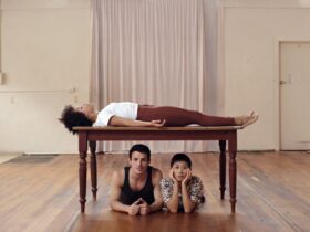 A person lies on a wooden table left to right. Under the table facing forward is a man and a woman.