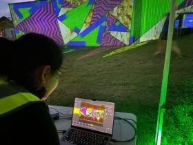 A person looking at a laptop screen which is projecting bright colours to a wall in the background