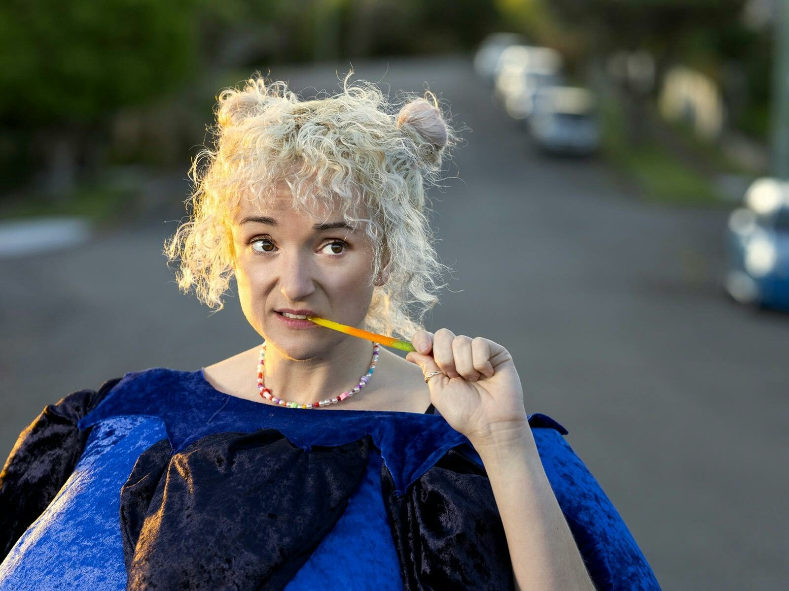 A young woman with blonde curly hair in 2 buns stands in a bluberry costume in the street.