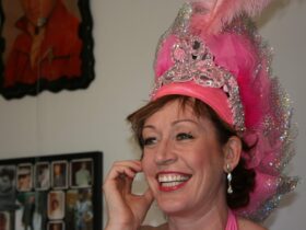 Headshot of performer Denise Hanlong, smiling and wearing a pink burlesque-style feather hat.