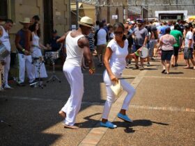 People dressed in white dancing outside with drummers at the side and public watching