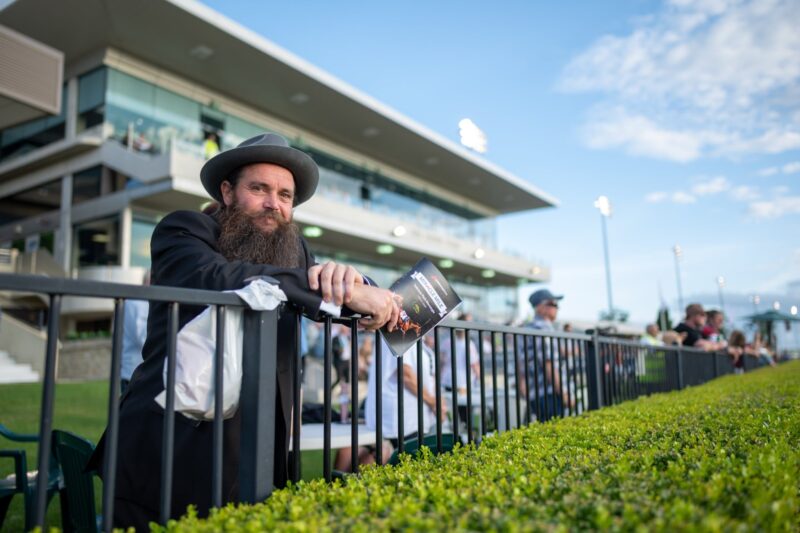 Man leaning on fence rail with Club Menangle grandstand in background on a sunny day